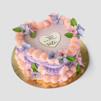 The Sugar Flower Heart Cake by Jenna Rae Cakes features pink and purple frosting, pastel flowers, green leaves, and a heart-shaped cookie with Happy 30th Birthday Tali, all presented on a gold board.