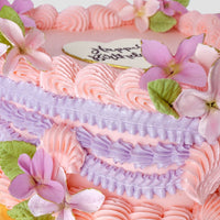The Sugar Flower Heart Cake by Jenna Rae Cakes is a heart-shaped cake with pink and purple buttercream swirls, topped with matching flowers and a white heart-shaped Happy Birthday plaque.