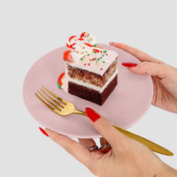 A person with red nails holds a pink plate with a slice of Jenna Rae Cakes’ Sugar Flower Sheet Cake, topped with white frosting, sprinkles, and candy, while holding a gold fork in the other hand.