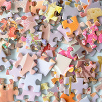 A close-up of scattered Sugar Rush Puzzle pieces by Jenna Rae Cakes, in pastel and vibrant colors, shows fragments of images on a light surface. The pieces are randomly arranged, not yet forming the finished picture.
