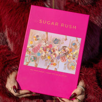 A person in a red faux fur coat holds a Jenna Rae Cakes Sugar Rush Puzzle box (500 pieces), which displays a vibrant dessert-themed table setting. Their nails are painted dark red.