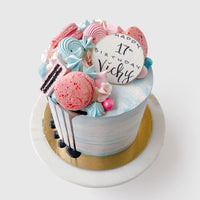 The Half Works Cake by Jenna Rae Cakes is a pastel birthday cake topped with pink and blue macarons, meringues, chocolate drizzle, and a white “Happy 17th Birthday Vicky” topper, served on a marble stand.