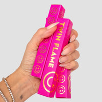 A hand with manicured nails holds two bright pink Twin Flame Perfume boxes from Jenna Rae Cakes, featuring gold lettering and smiley face designs; the wearer has a beaded bracelet and a small wrist tattoo.