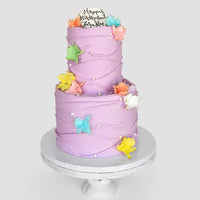 The Waves and Pearls cake by Jenna Rae Cakes is a two-tiered lavender creation adorned with colorful edible butterflies, white pearls, and a “Happy Birthday Zariah” topper, presented on a white cake stand.