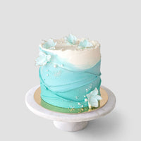 The Waves and Pearls cake by Jenna Rae Cakes features blue and white ombre textured icing, adorned with edible blue and white flowers and pearl-like sprinkles, displayed on a marble stand against a light gray background.