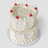 The “Wedding Cakes Under $500” by Jenna Rae Cakes is a two-tiered white frosted cake with piped cream designs, six red cherries on top, and cursive writing on the upper tier, presented on a white cake board.