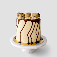 The Cookie Dough Cake by Jenna Rae Cakes features smooth beige frosting, chocolate drips down the sides, and scoops of cookie dough on top, displayed on a marble cake stand against a plain background.