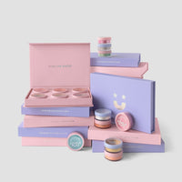 Pastel pink and lavender Dazzle Dust Boxes and jars by Jenna Rae Cakes, some open to reveal shimmer powders. The minimalist packaging features the phrase MAKE LIFE DAZZLE.