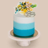 A custom blue ombre “Design Your Own Cake” by Jenna Rae Cakes sits on a marble stand, topped with cartoon animals and a “Happy Birthday Leo” topper.