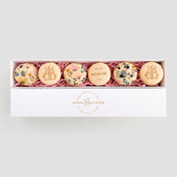 A white box labeled Jenna Rae Cakes contains six Favours macarons with floral designs and text, nestled in pink shredded paper. The center macaron features THANK YOU 10.15.22.