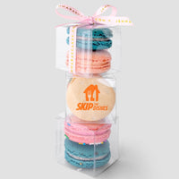 A stack of three clear Jenna Rae Cakes Favours boxes, each filled with colorful macarons and tied with a pink ribbon. The middle box features a macaron printed with the SkipTheDishes logo.