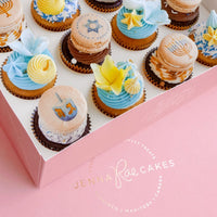 Hanukkah Works Cupcakes by Jenna Rae Cakes: a box of Hanukkah-themed cupcakes and macarons with blue, yellow, and white frosting, decorated with stars, menorahs, and dreidels. Logo displayed on a pink surface.