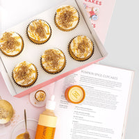 A box of six pumpkin spice cupcakes with swirled frosting sits beside the open Jenna Rae Cakes and Sweet Treats Cookbook, a piping bottle, a small container of sprinkles, and a bowl with cupcake crumbs.