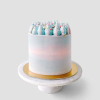 The Last Minute Cakes by Jenna Rae Cakes features a tall, round cake with smooth pastel blue and pink frosting, topped with blue and pink swirled dollops and colorful sprinkles on a white marble cake stand.
