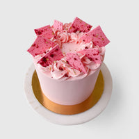 The Lemon Raspberry Cakes 101 In-Person Cake Decorating Class by Shop Jenna Rae Cakes features a round pink cake adorned with rosettes, chocolate shards, and gold sprinkles, displayed on a white marble cake stand with a gold base.