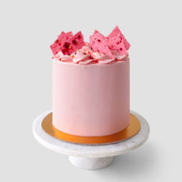 The Lemon Raspberry Cakes 101 In-Person Cake Decorating Class by Shop Jenna Rae Cakes features a round pink frosted cake with decorative shards and sprinkles, displayed on a white marble stand with a gold base against a light gray background.