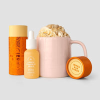 A pink mug with whipped cream sits beside orange and yellow containers and a Jenna Rae Cakes Liquid Pumpkin Spice dropper bottle, all set against a light gray background.