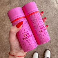 A hand with red-painted nails holds two pink Jenna Rae Cakes Liquid Red Velvet bottles against a brown textured background. White shoes and part of a bracelet are visible at the bottom of the image.
