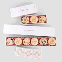 Three white Favours boxes from Jenna Rae Cakes, each holding decorated macarons with floral designs and gold initials, displayed on a light gray background. One macaron features THANK YOU 10 25 22.