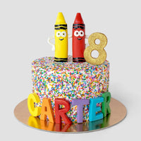 A colorful birthday cake covered in rainbow sprinkles, topped with two crayon figures, a glittery gold number 8, and the name CARTER spelled out in large, colorful letters at the base.