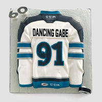 A cake shaped like a white and teal hockey jersey with the name DANCING GABE and the number 91. The cake also features a fondant hockey puck and the number 60 on the corner.