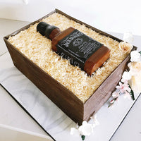 A realistic cake designed to look like a wooden crate filled with shredded paper, containing a bottle resembling Jack Daniel’s whiskey, decorated with white and pale pink flowers.