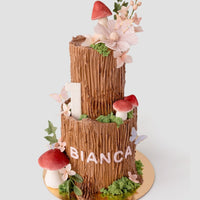 A two-tiered cake decorated to look like tree trunks, with red and white mushrooms, green moss, pastel flowers, butterflies, a number 1 candle, and the name Bianca in white letters on the lower tier.