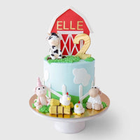 A farm-themed birthday cake with a red barn topper reading ELLE, a gold number 2, and fondant animals including a cow, pig, sheep, and chickens, with clouds and grass decorations.
