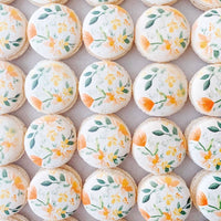 Neatly arranged on a light surface, Jenna Rae Cakes Printed Macarons feature white tops adorned with hand-painted orange flowers and green leaves.
