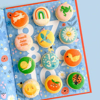 Jenna Rae Cakes Printed Macarons, featuring cute designs like rabbits, rainbows, and ladybugs, are beautifully arranged on an open book with a blue floral and ladybug pattern.