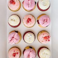 Enjoy a box of twelve Regular Cupcakes from Jenna Rae Cakes, each topped with pink, white, and purple swirled frosting, sprinkles, and finished with dried fruit or chocolate pieces.