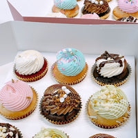 An open box and tray of Jenna Rae Cakes Regular Cupcakes, each topped with colorful swirled frosting, sprinkles, and decorative toppings, arranged neatly in rows.