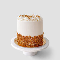 The Salted Caramel Crunch Cake by Jenna Rae Cakes is a round, white frosted cake with whipped cream swirls and crushed nuts around the base, displayed on a marble stand against a plain light background.