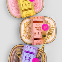 Three packaged Snack Cakes by Jenna Rae Cakes—pink, caramel, and chocolate—each with decorative labels and a gold fork on top, arranged on a light grey background.