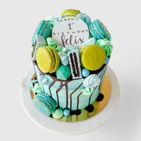The Full Works Cake by Jenna Rae Cakes is a tall blue and green birthday cake topped with macarons, meringues, chocolates, and a large cookie reading “Happy 1st Birthday Felix,” finished with chocolate drips on a marble plate.