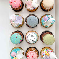 The Works Cupcakes by Jenna Rae Cakes features 12 uniquely decorated cupcakes with colorful frosting, sprinkles, macarons, flowers, and edible pearls, neatly arranged in three rows of four.