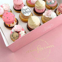 The Works Cupcakes by Jenna Rae Cakes come in a pink box with a gold logo and include assorted cupcakes topped with pink and gold frosting, macarons, cookie dough, meringues, and a “Happy Birthday” round topper.
