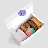 The Jenna Rae Cakes Sweet Treat Gift Box includes two large cookies, a pink and blue round treat, a small tub of frosting, and a macaron printed with two women smiling.