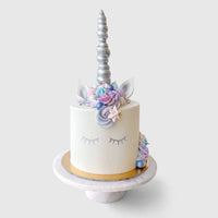 Join the Unicorn In-Person Cake Decorating Class by Shop Jenna Rae Cakes to create a white unicorn cake with closed eyes, silver horn, pastel floral mane, and ears, all beautifully displayed on a marble stand against a simple backdrop.