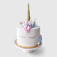 The Unicorn Cake by Jenna Rae Cakes features white frosting, a gold horn, closed eyes, pastel flowers, and ears on top, displayed on a marble cake stand against a plain background.