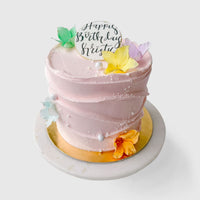 The Waves and Pearls cake by Jenna Rae Cakes features pink frosting, colorful edible butterflies, white sprinkles, a round Happy Birthday topper, and sits on a gold and white cake board.