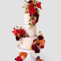 A two-tiered white cake on a stand, decorated with vibrant red and orange flowers, gold leaves, and assorted fresh fruits like figs, grapes, and berries, against a plain light gray background.