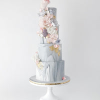 A four-tiered marble-patterned cake with gold accents, decorated with pastel pink, white, and purple sugar flowers cascading down one side, displayed on a white cake stand with a gold rim.