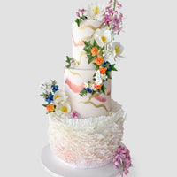 A three-tiered white cake decorated with pink and gold brush strokes, adorned with colorful, realistic sugar flowers and intricate white ruffles around the base. The cake sits on a plain white round board.