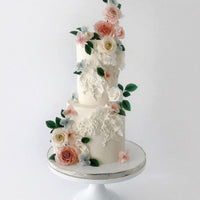 The Jenna Rae Cakes Wedding Cakes Under $1,000 collection features elegant two-tiered white cakes, adorned with realistic pink, white, and peach sugar flowers with green leaves, beautifully displayed on a simple white cake stand.