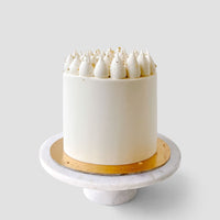 A Wedding Cake from Jenna Rae Cakes under $200 features white frosting, whipped cream dollops, and gold sprinkles on a gold cake board and white marble stand against a light gray background.