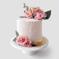 The Jenna Rae Cakes Wedding Cakes Under $200 features a white frosted cake on a marble stand, adorned with pink and peach roses and green leaves.