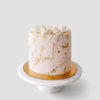 The “Wedding Cakes Under $200” by Jenna Rae Cakes features a round white frosted cake with gold splatter, white rosettes, on a gold board and marble stand, set against a light gray background.