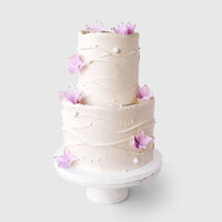 A two-tiered white frosted cake with light pink edible flowers and pearls by Jenna Rae Cakes, featured in Wedding Cakes Under $500, displayed on a white stand against a plain light background.