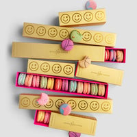 Jenna Rae Cakes’ $100 JRC Gift Bag features gold boxes with smiley faces filled with pink, blue, green, and yellow macarons, artfully arranged on a light gray surface with some treats scattered outside.